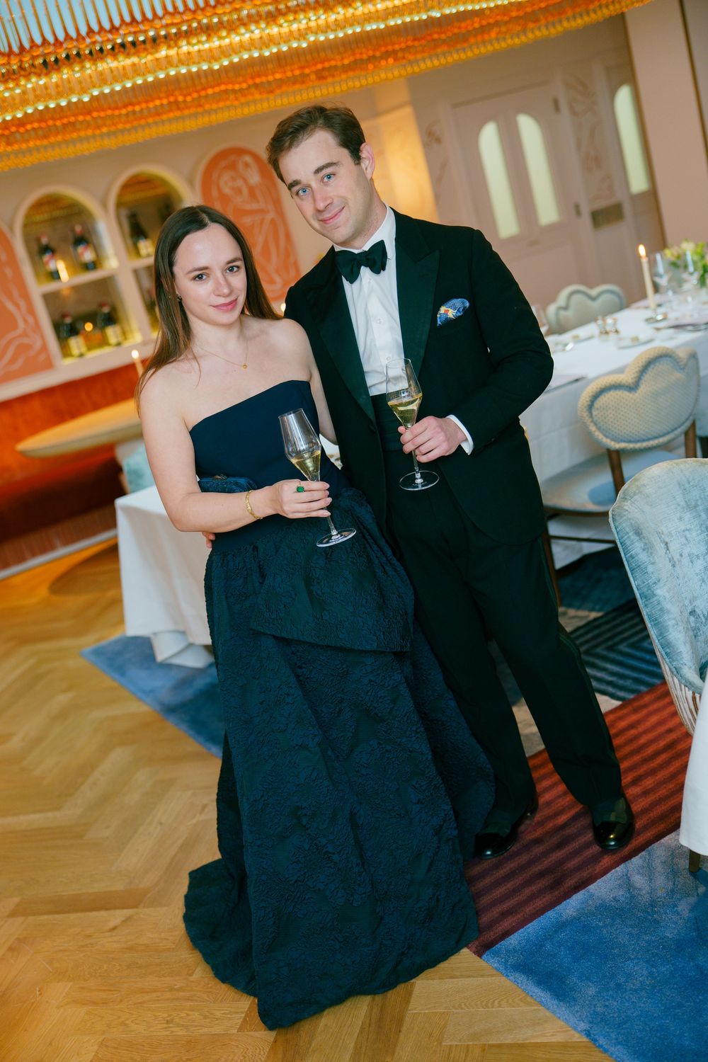 Elegant couple in formal attire holding champagne glasses at a upscale event, woman in navy gown, man in black tuxedo.