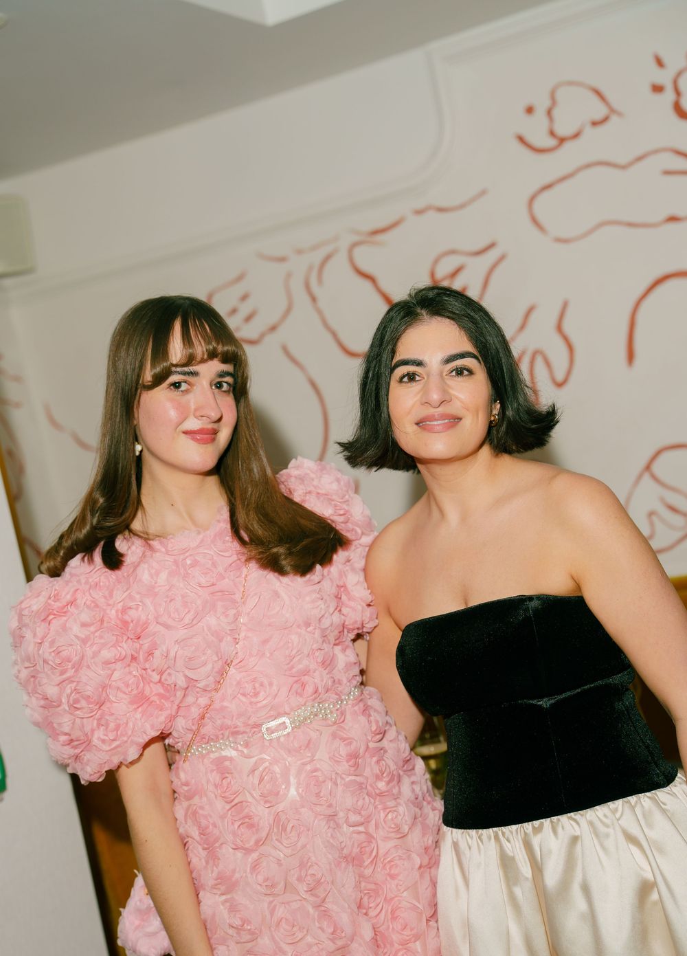 Two young women posing together, one in a pink floral dress and one in a black and white strapless gown.