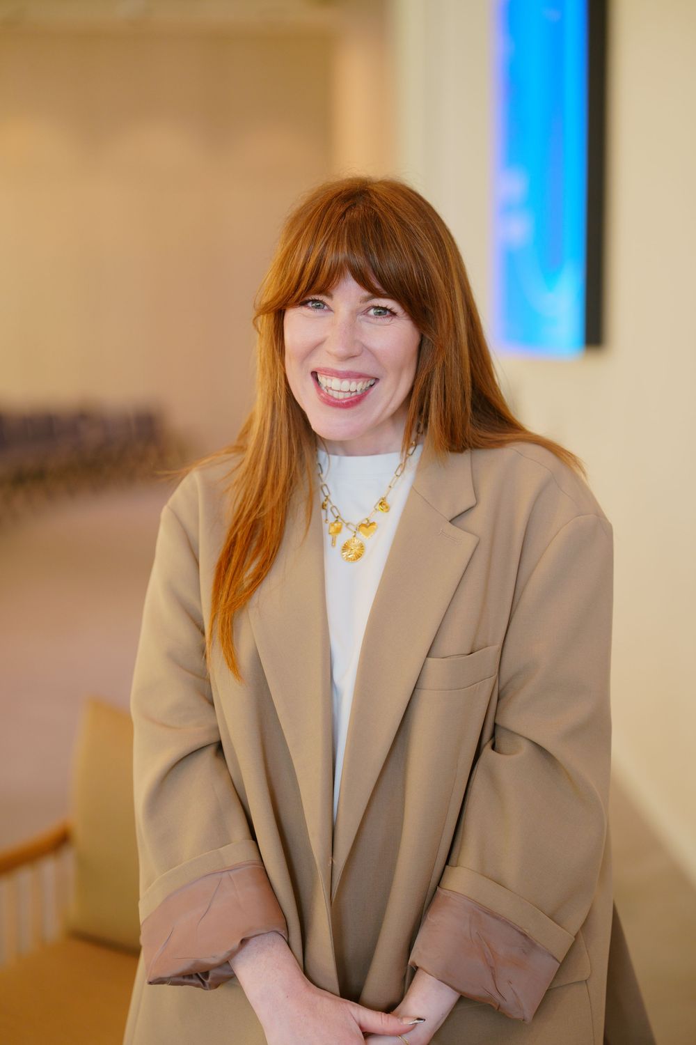 Smiling woman with red hair wearing a tan blazer and gold necklace stands in a bright modern interior space.
