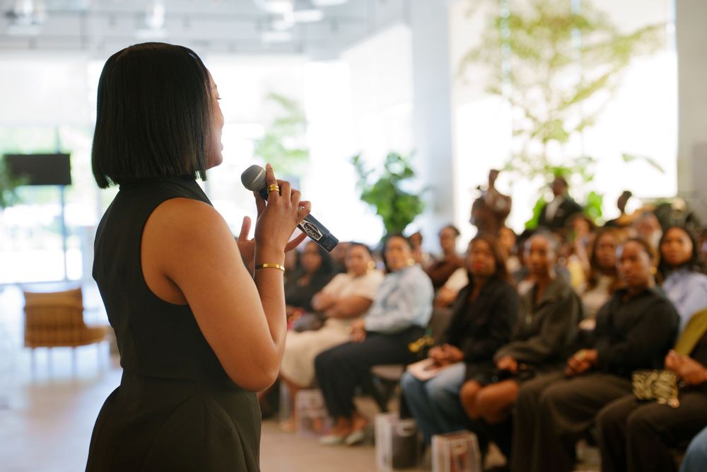 Woman speaker addresses diverse audience at indoor event, holding microphone in modern bright venue.