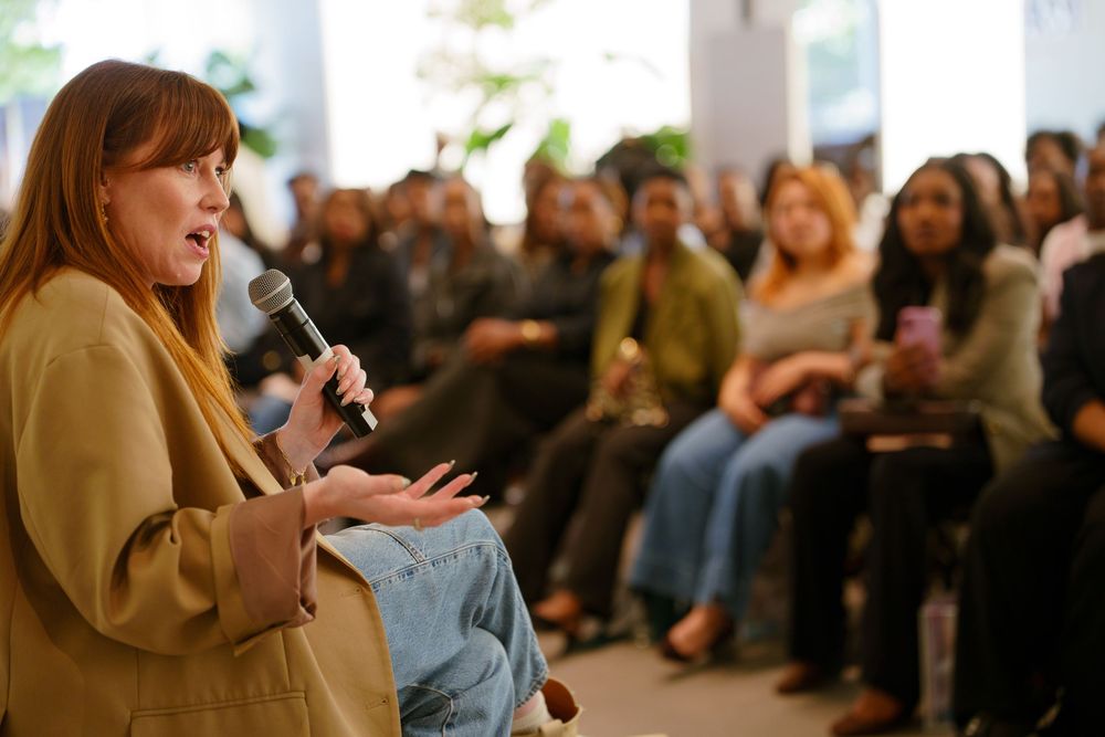 Woman speaking into microphone at indoor event with diverse audience seated in rows behind her.