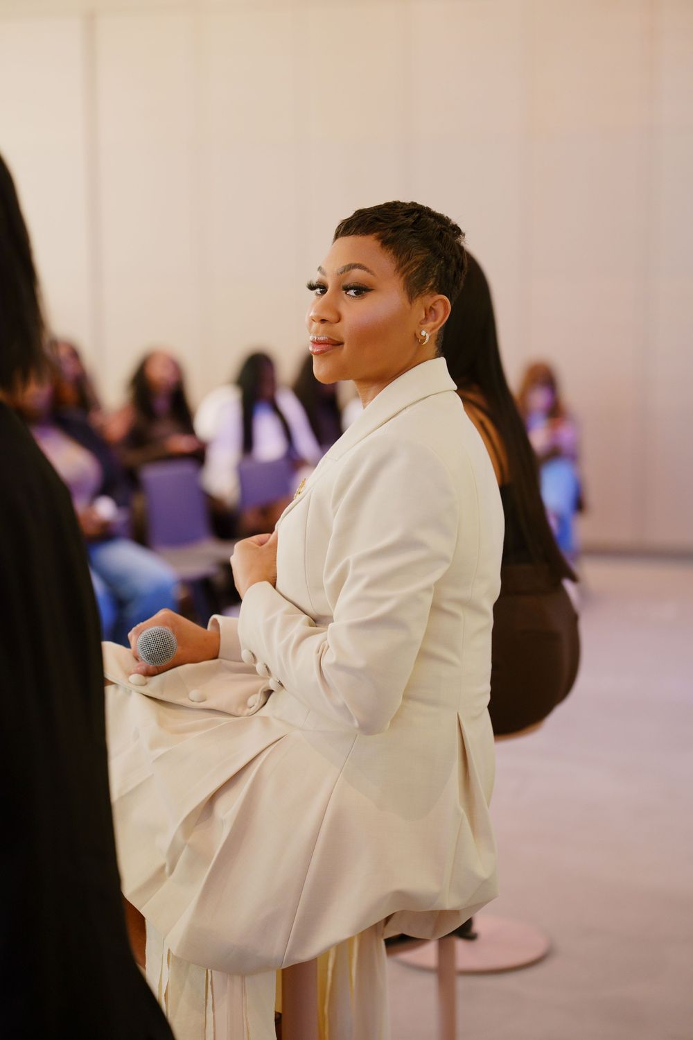 Woman in white blazer sits confidently at an indoor event, smiling softly with braided hair and gold earrings.