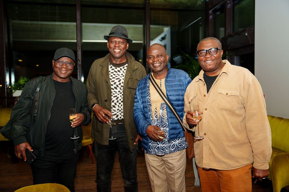 Four people stand together smiling at a social gathering, wearing casual attire including hats and jackets.