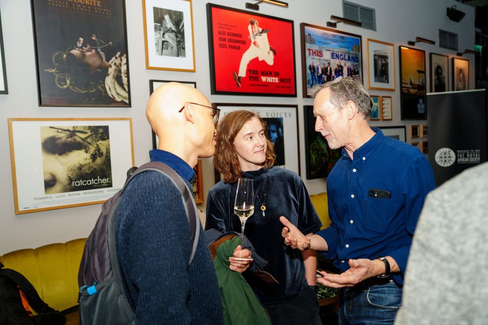 A casual social gathering at an art gallery with people chatting while surrounded by framed artwork on the walls.
