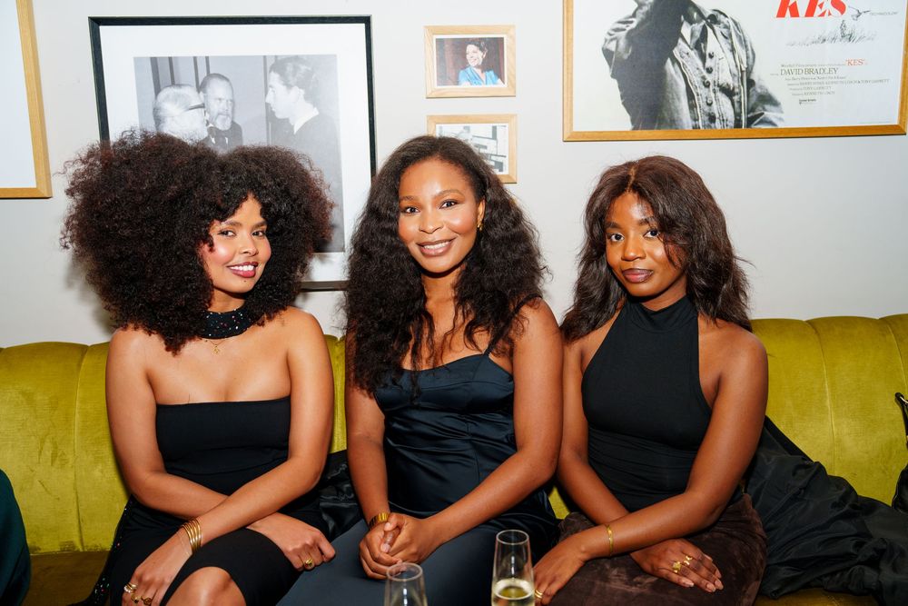 Three women in elegant black dresses sit together on a yellow-green couch at a social gathering.