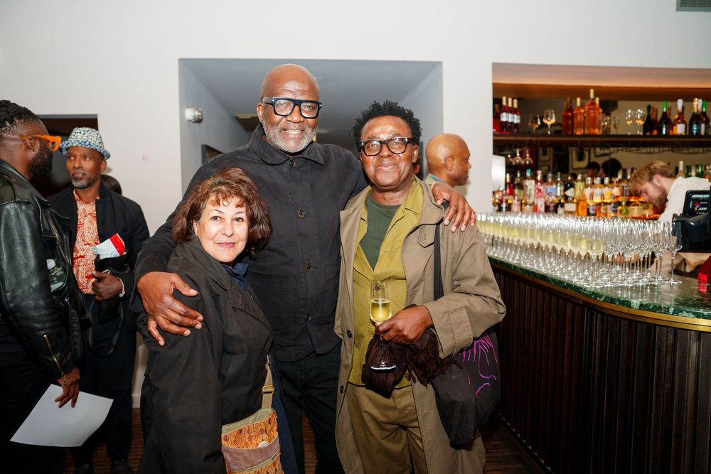 Three friends gather together for a photo at a bar with glassware and bottles visible in the background.