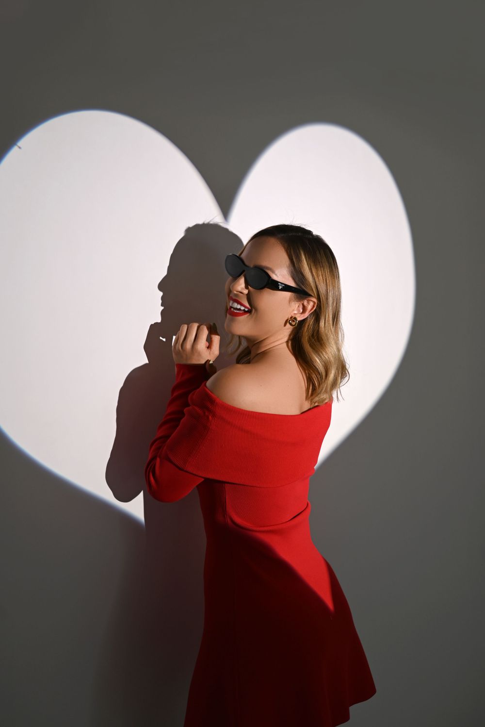 A figure in a red dress poses against a white heart-shaped background casting dramatic shadows.