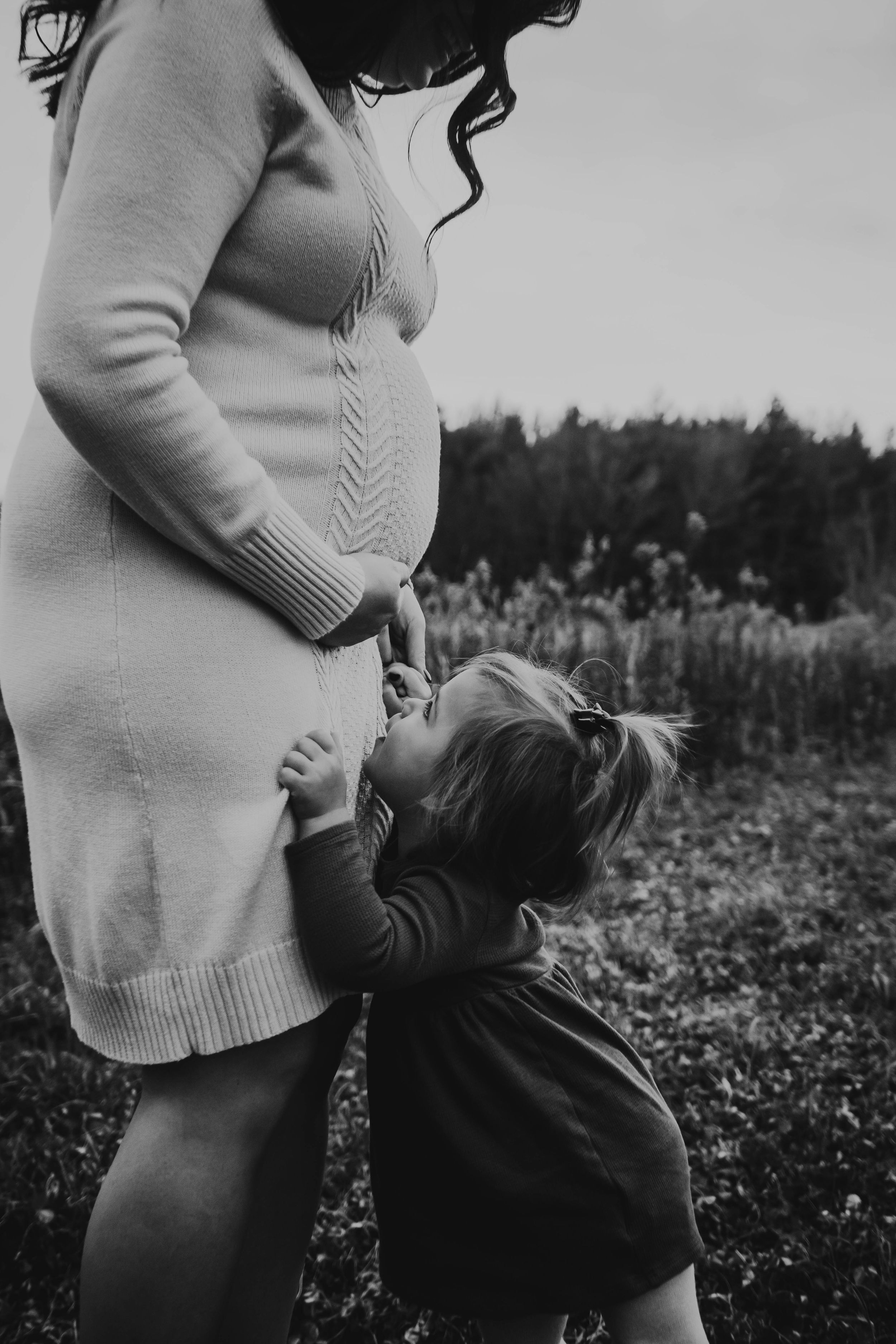 Black and white photo showing pregnant woman in sweater with small child touching her belly outdoors in nature.