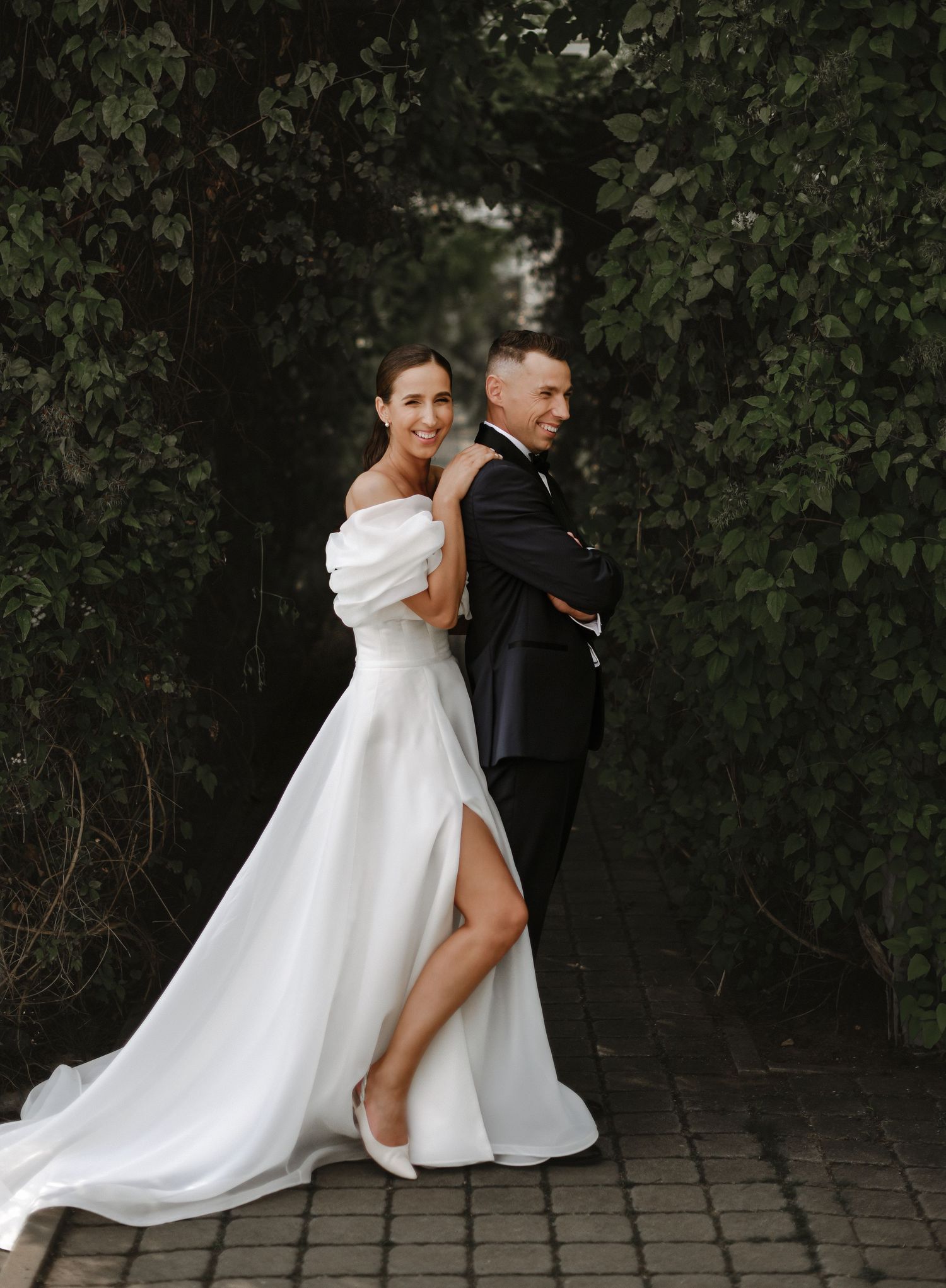 Artistic wedding portrait of couple posing back-to-back against greenery.