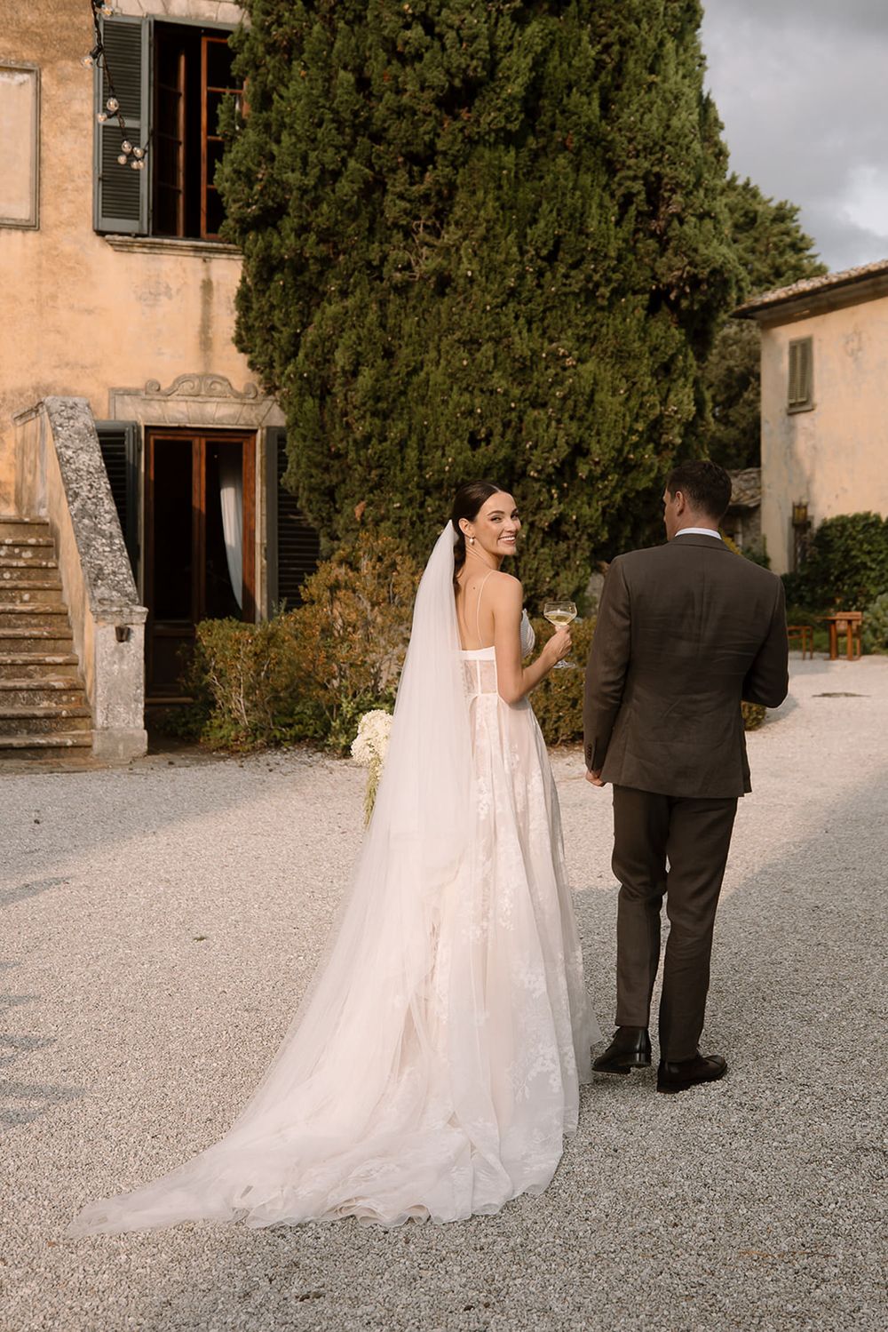A wedding couple in formal attire standing on a gravel path outside a rustic Italian villa.