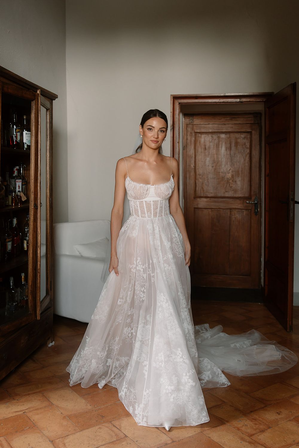 Elegant white corset wedding gown with lace overlay and flowing train displayed in a rustic room.