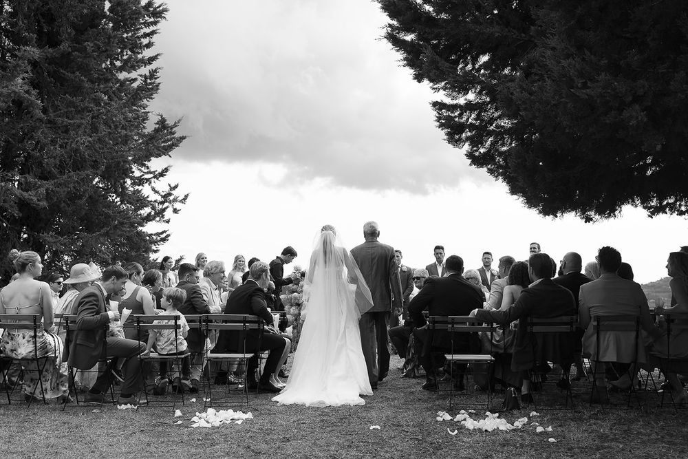 Outdoor wedding ceremony with guests seated beneath tall pine trees overlooking a scenic view.
