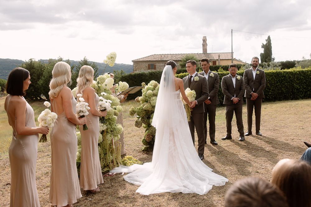 Wedding party lined up outdoors with bridesmaids in beige dresses and groomsmen in dark suits.
