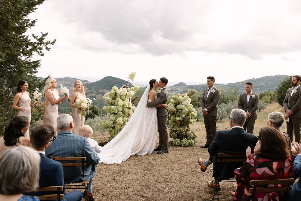 Outdoor mountain wedding ceremony with guests seated on white chairs among evergreen trees.