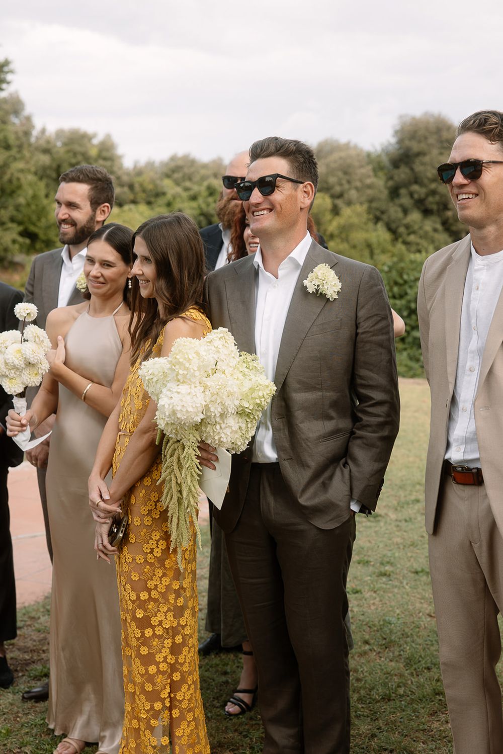 Wedding party in formal attire stands together holding bouquets at outdoor ceremony.