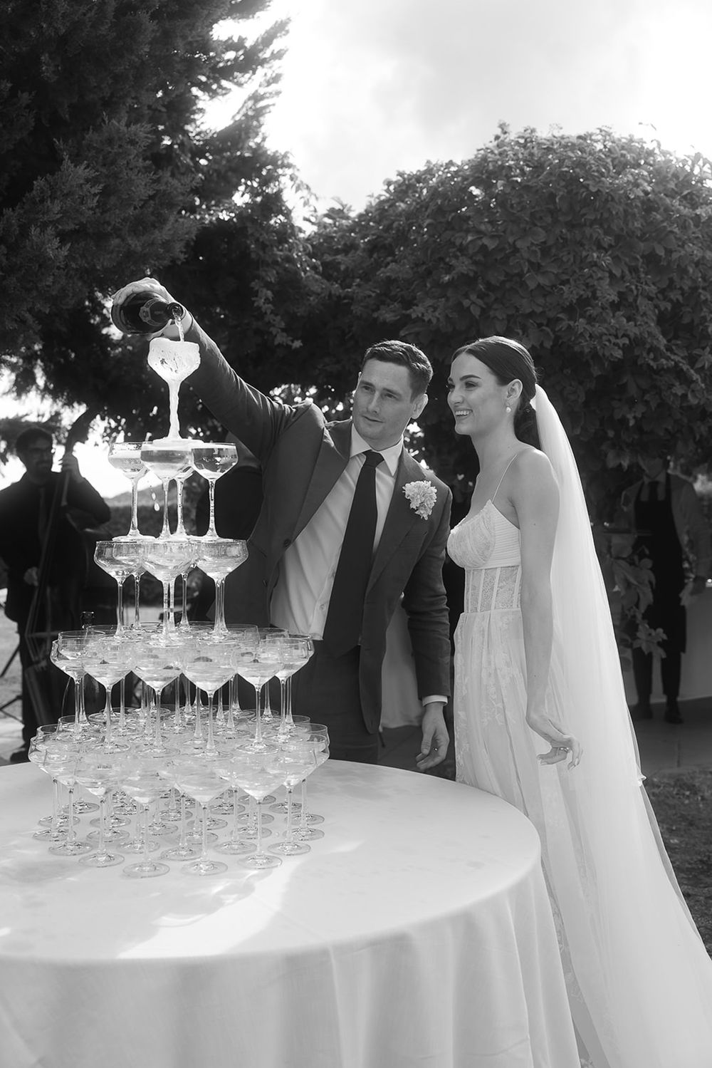 Black and white photo of couple pouring champagne into tower of glasses at outdoor reception.