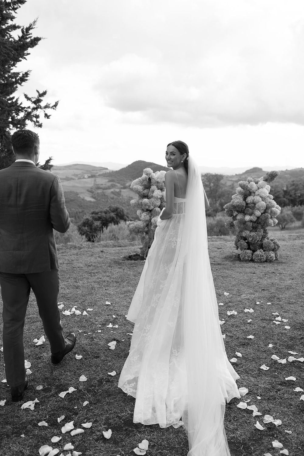 Scenic black and white wedding photo with rolling hills and floral arrangements in background.