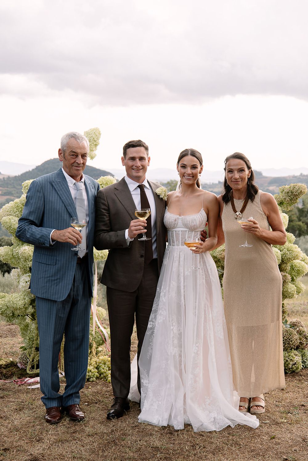 Group poses with drinks against scenic hillside backdrop at formal outdoor celebration.
