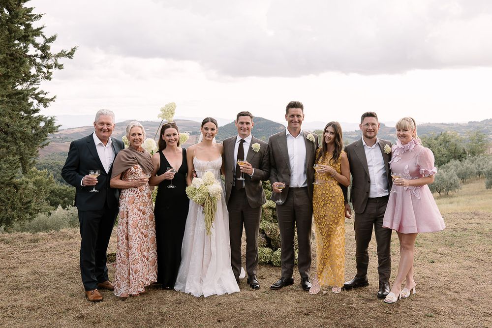 Wedding party group photo with guests in colorful formal attire against rural landscape.