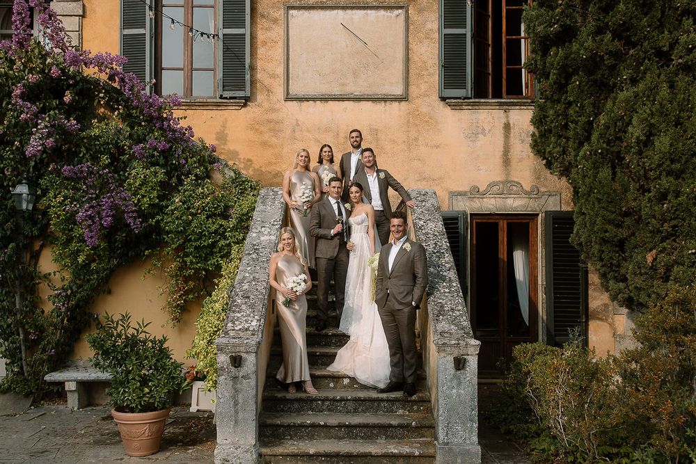 Wedding party posed on stone steps of Italian villa with greenery and cream colored walls at sunset.