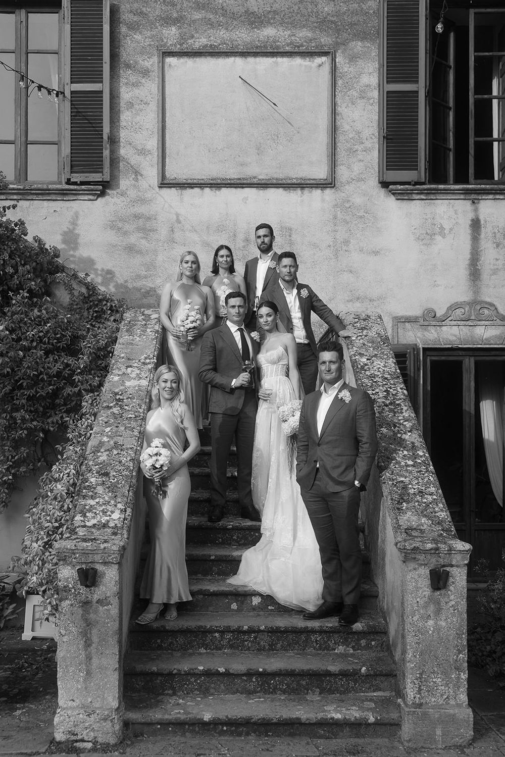 Black and white photo of wedding party gathered on historic stone staircase against textured villa wall.