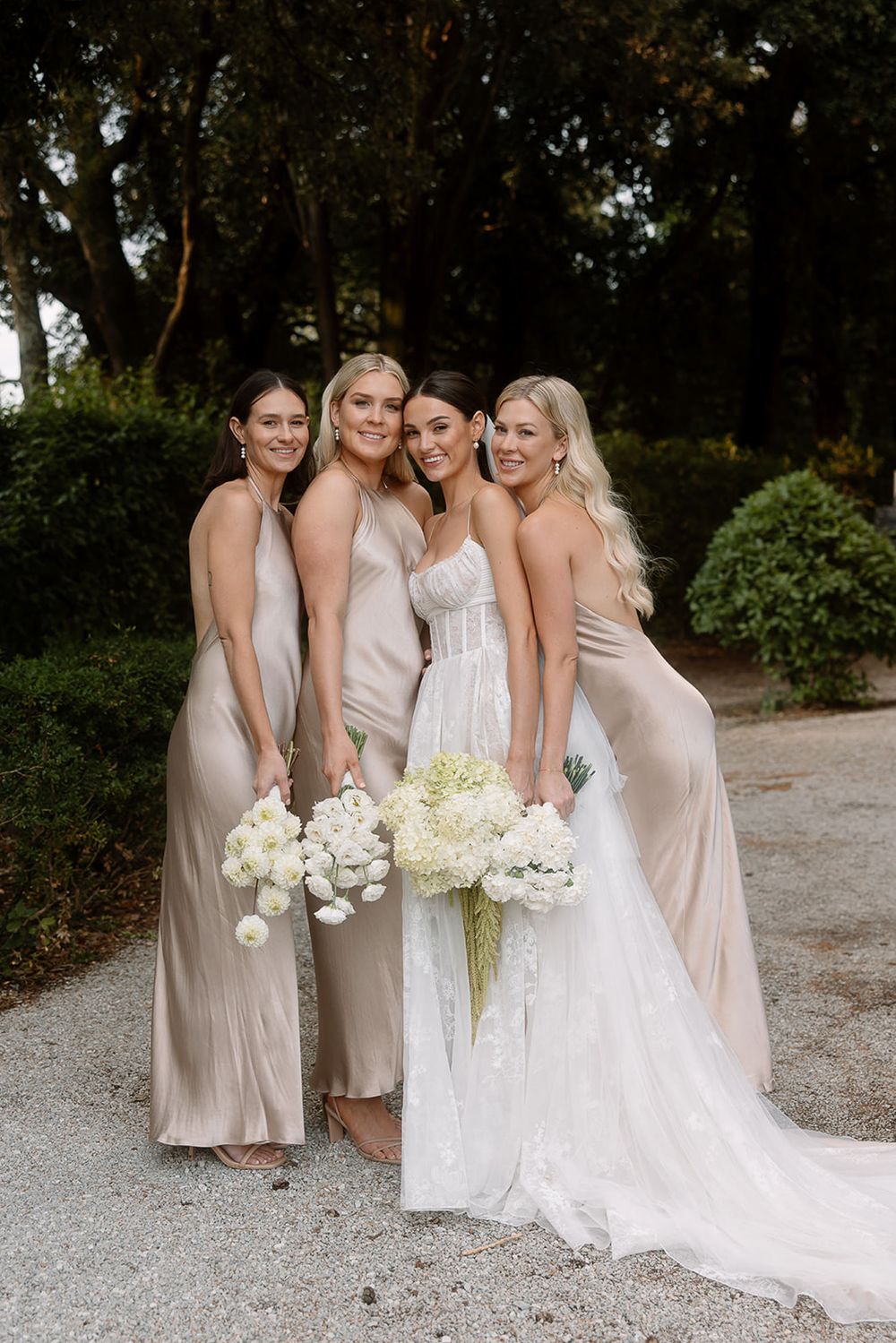 Bridal party in blush gowns holding white bouquets pose together on garden path.