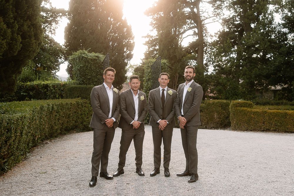 Groomsmen in grey suits pose on gravel path with garden hedges at golden hour.