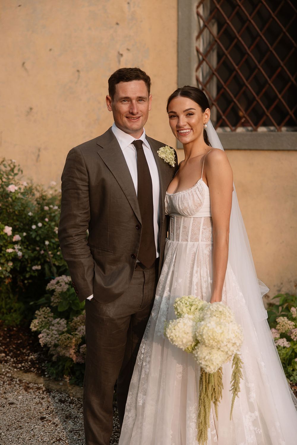 Wedding couple poses against cream villa wall with climbing roses and rustic window.