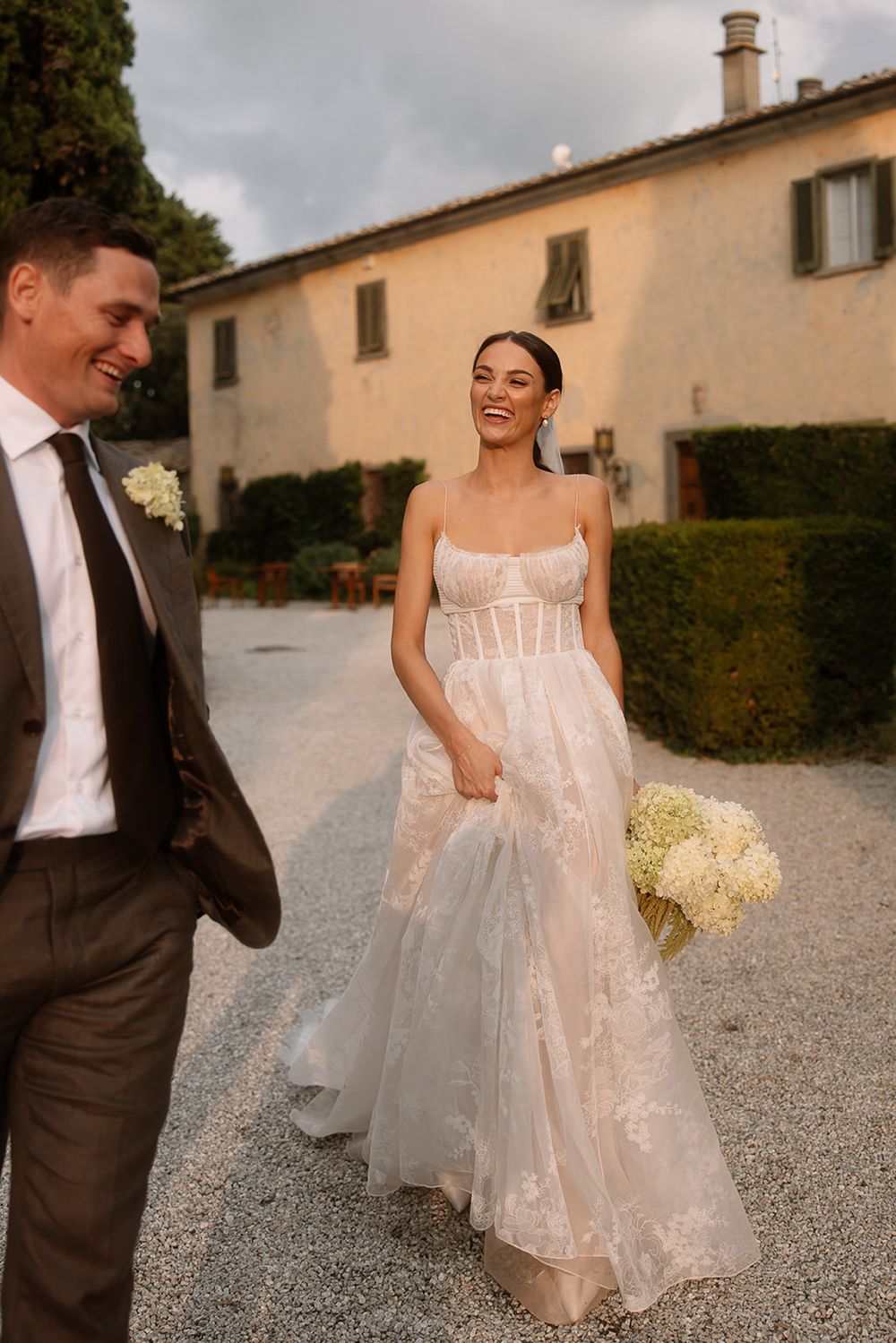 Couple shares candid moment on gravel path with Italian villa in background at sunset.