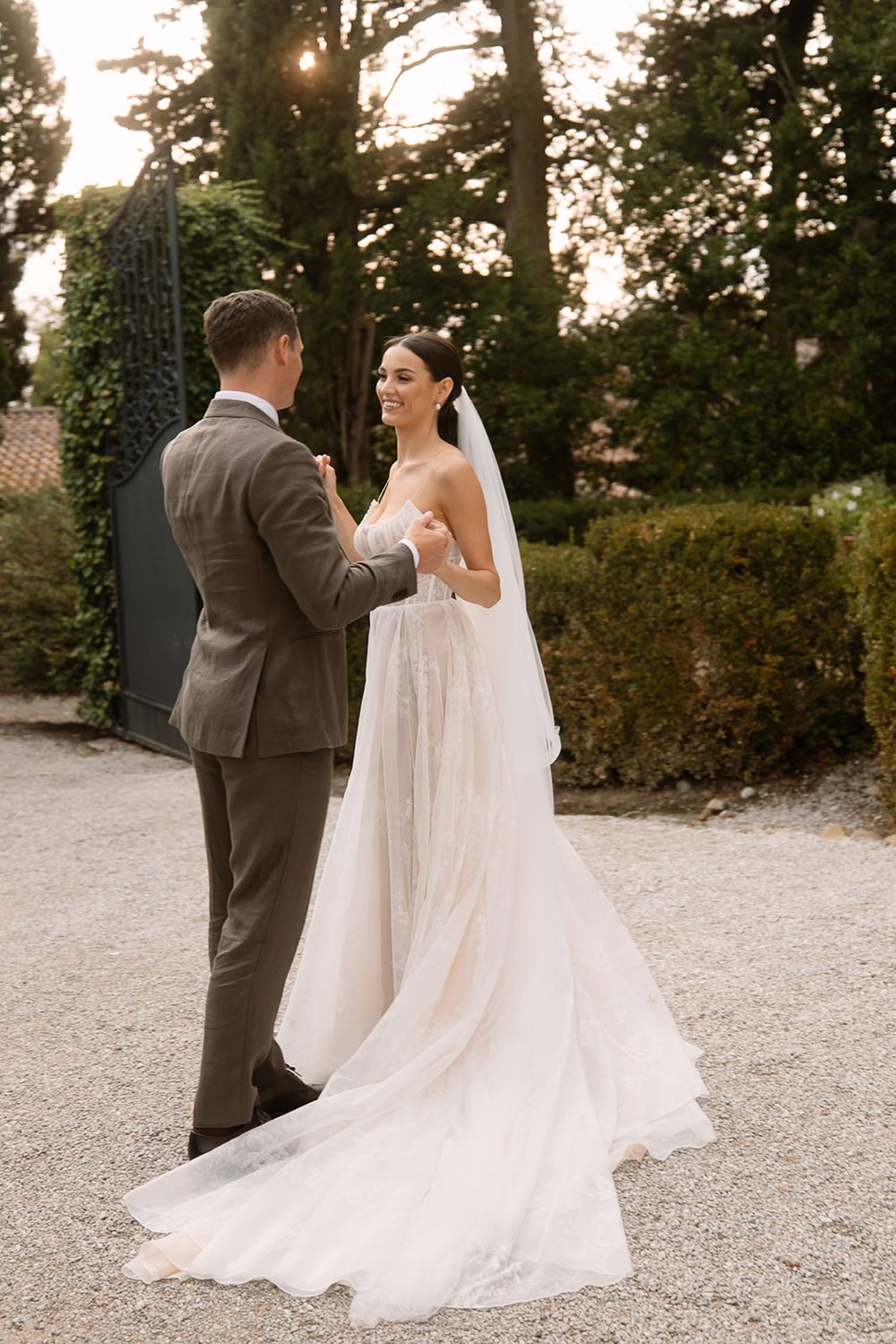 Elegant couple in wedding attire sharing intimate moment on garden path with long veil trailing.