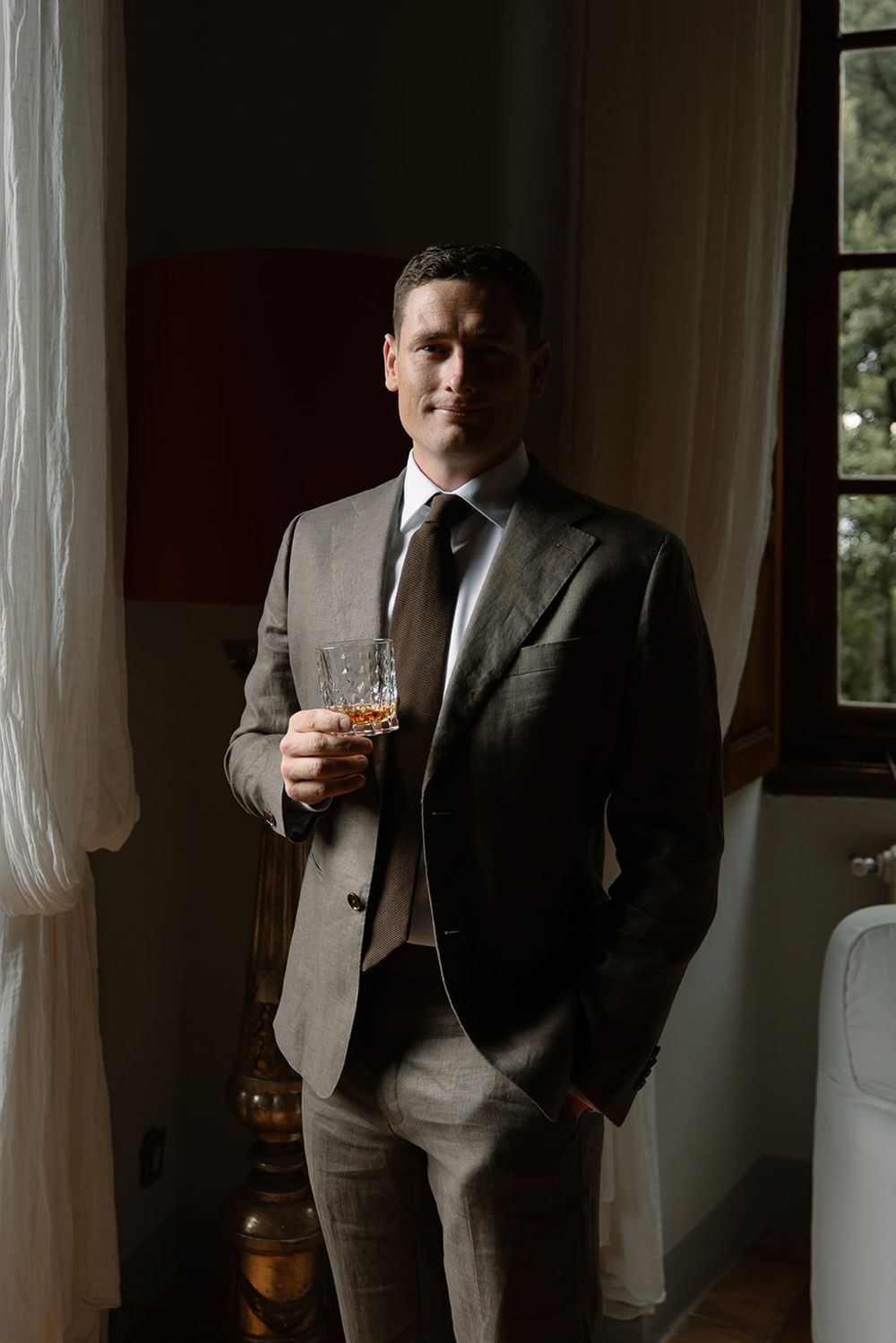 Elegant groom in grey suit holding whiskey glass by window.