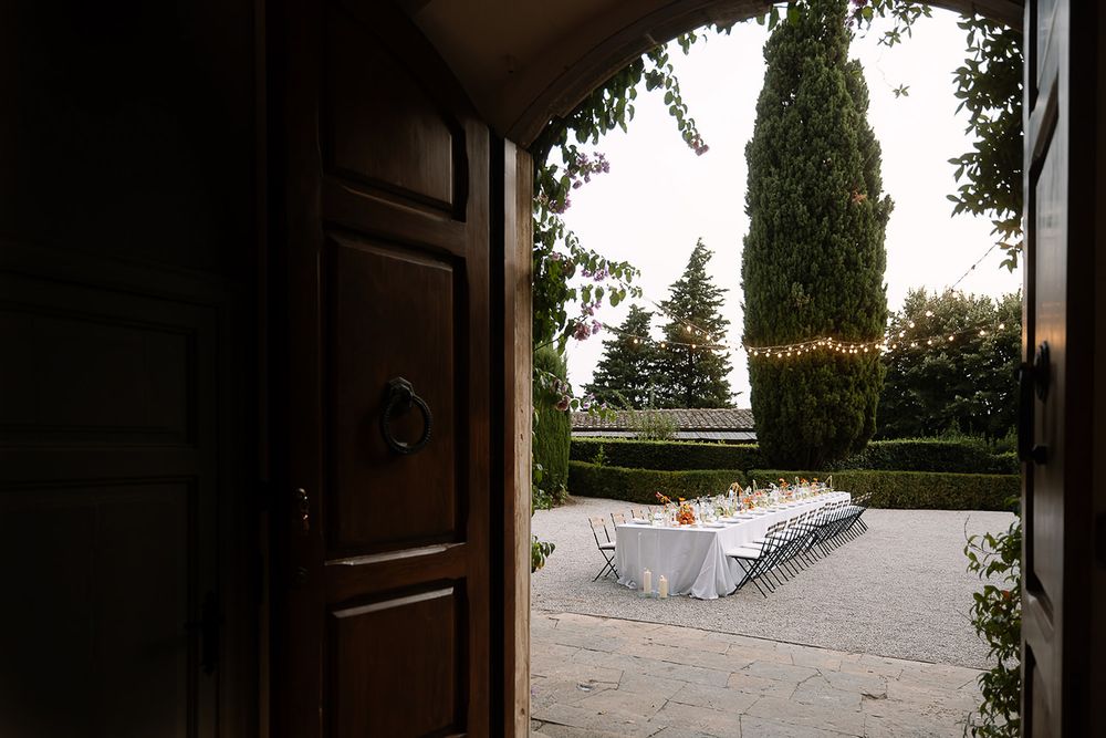 View through an open wooden door to an elegant outdoor dining setup with white tablecloths and cypress trees.