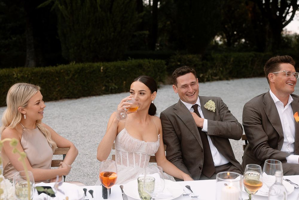Wedding party seated at an outdoor dining table sharing drinks and laughter.
