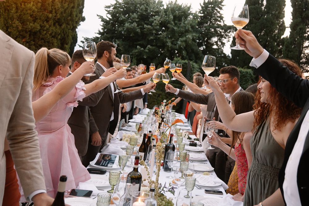 Wedding guests raising glasses in a toast across a long outdoor dining table.