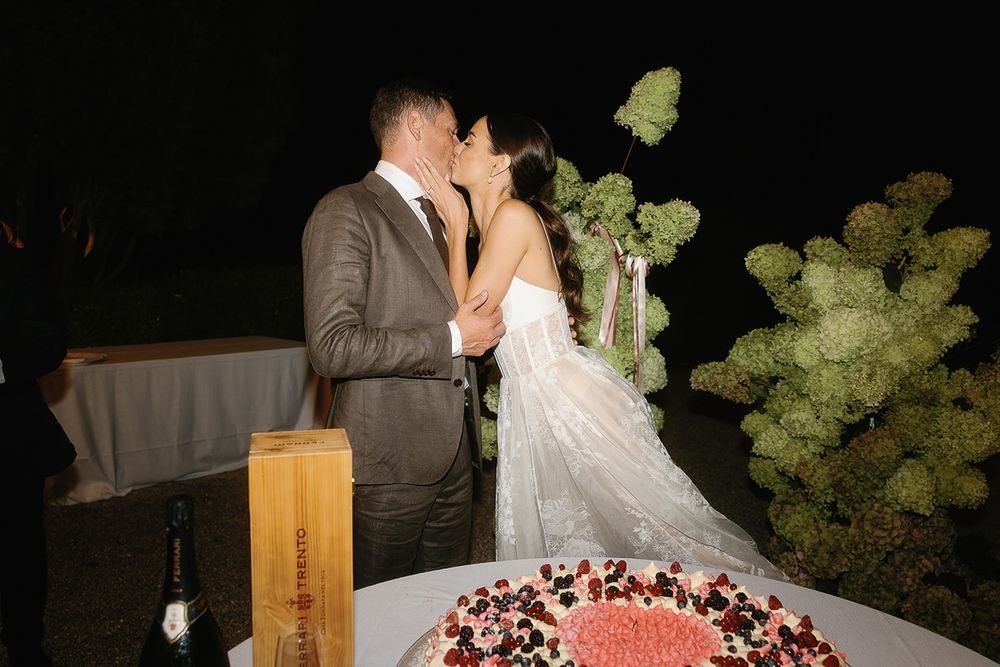 Newlyweds sharing a romantic kiss beside their wedding cake at a nighttime reception.