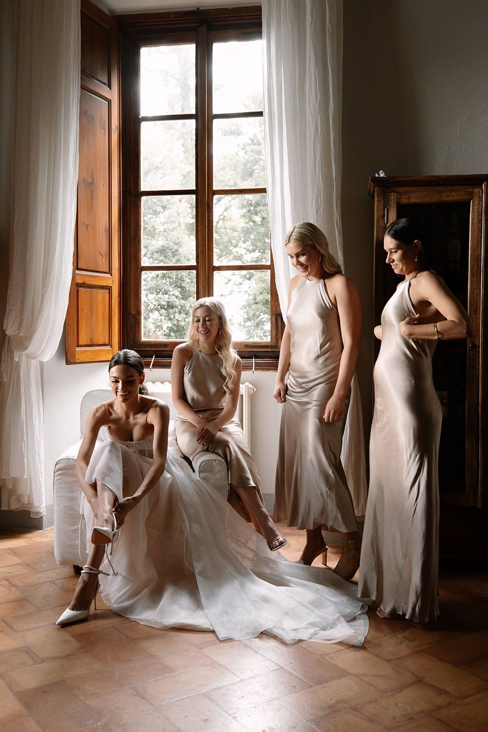 Wedding party in elegant silk dresses gathered by sunlit window.