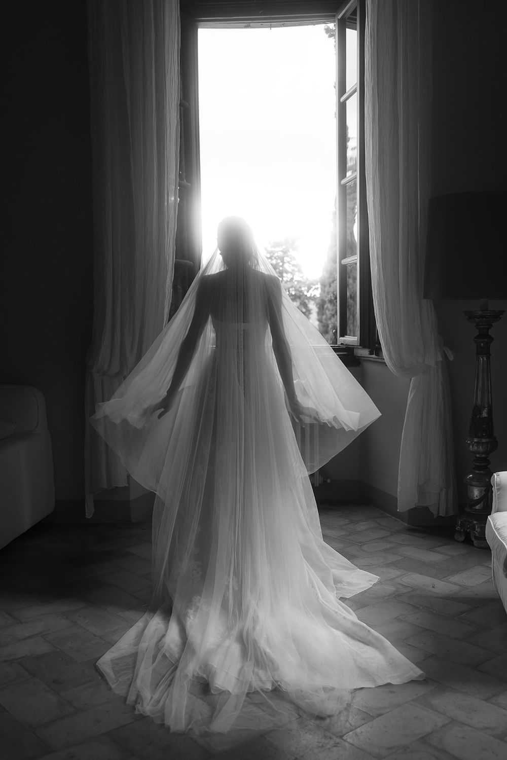 Silhouette of bride in flowing dress against bright window in black and white.