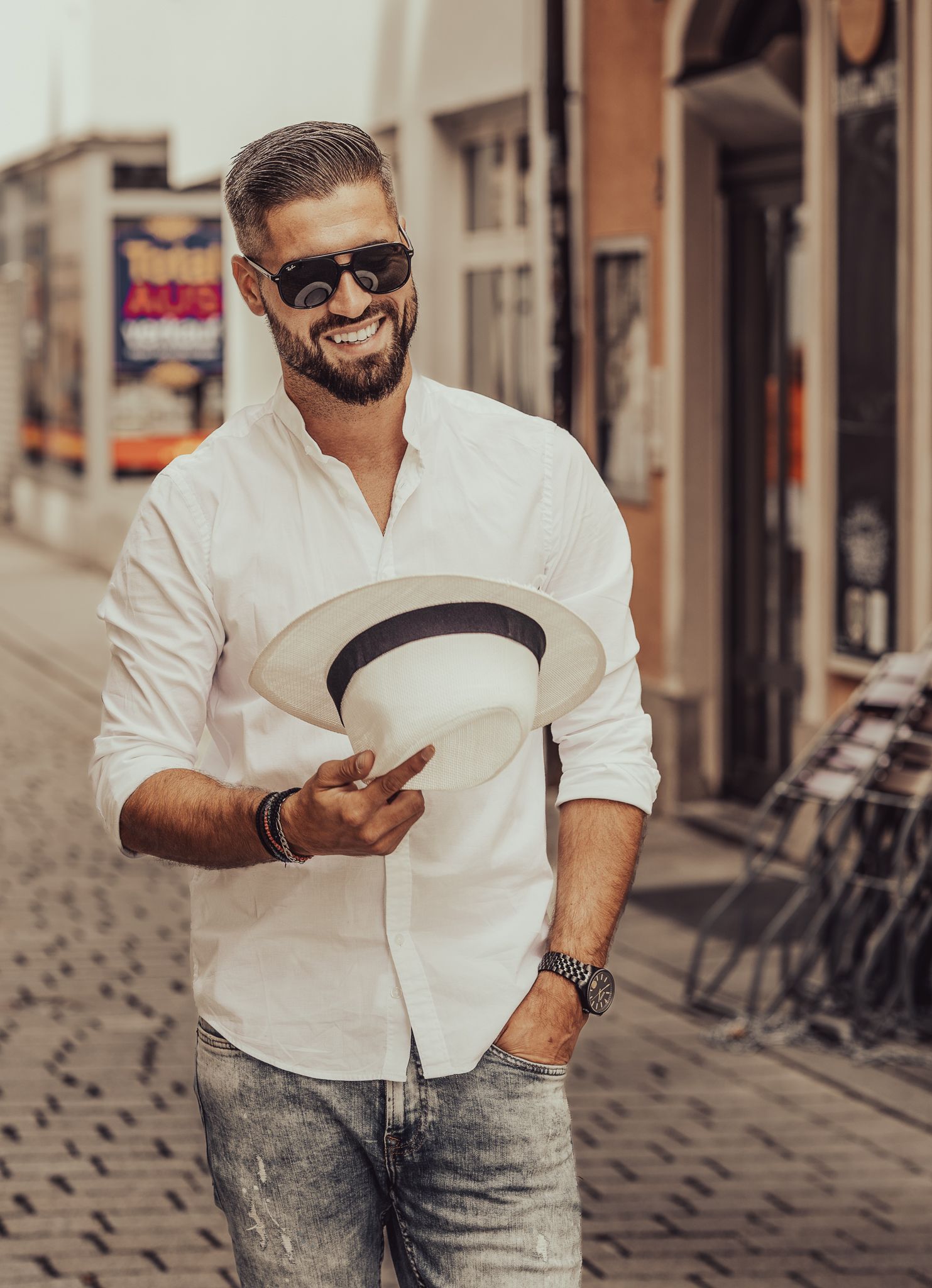 Menfotografie, Fashionfotografie, Stylish man in white shirt and sunglasses holding a hat while standing on a city street.