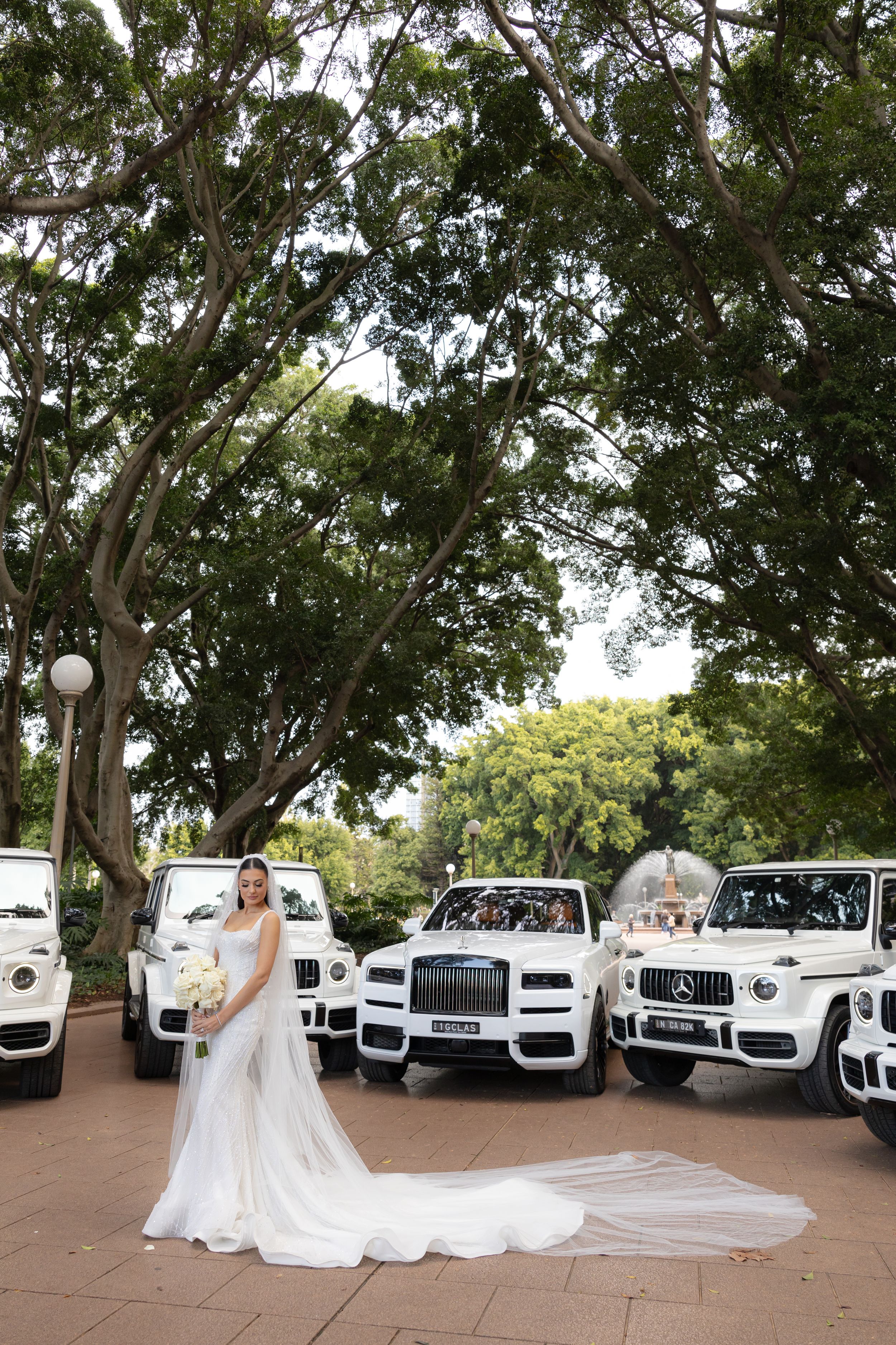 Bride in a stunning white gown posing before a fleet of luxury white SUVs under lush trees.