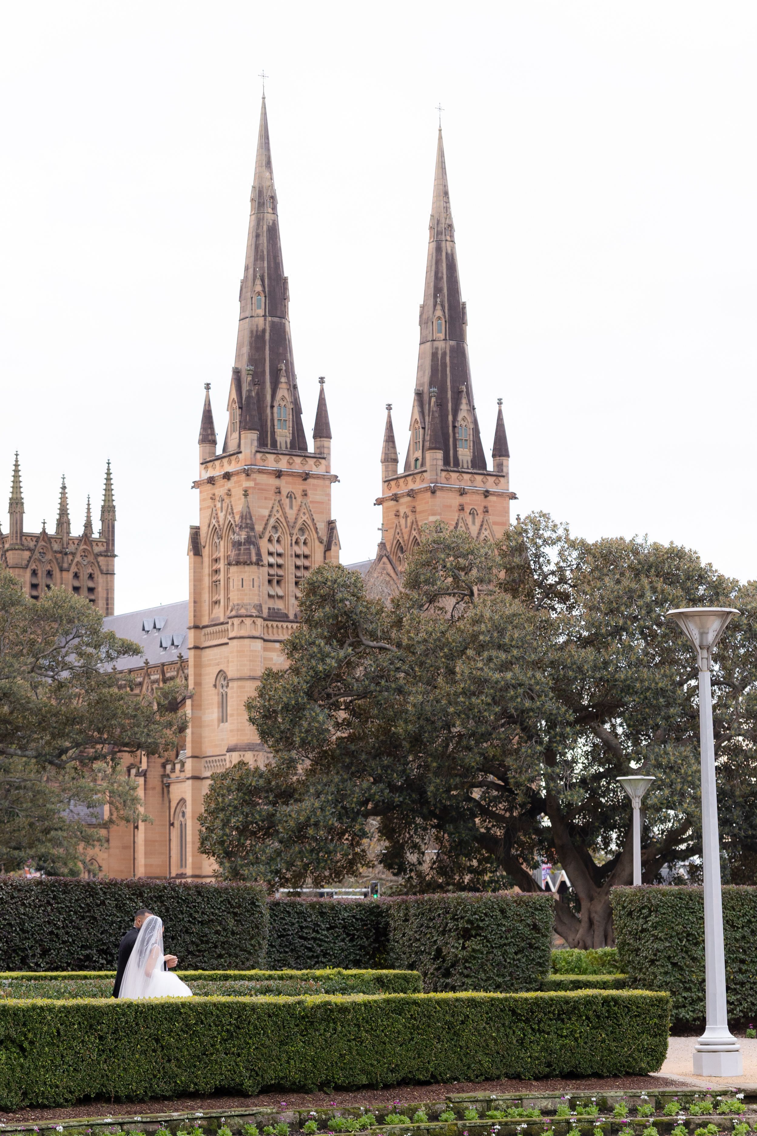 Bride in white gown walking in a manicured garden with Gothic cathedral spires in background.