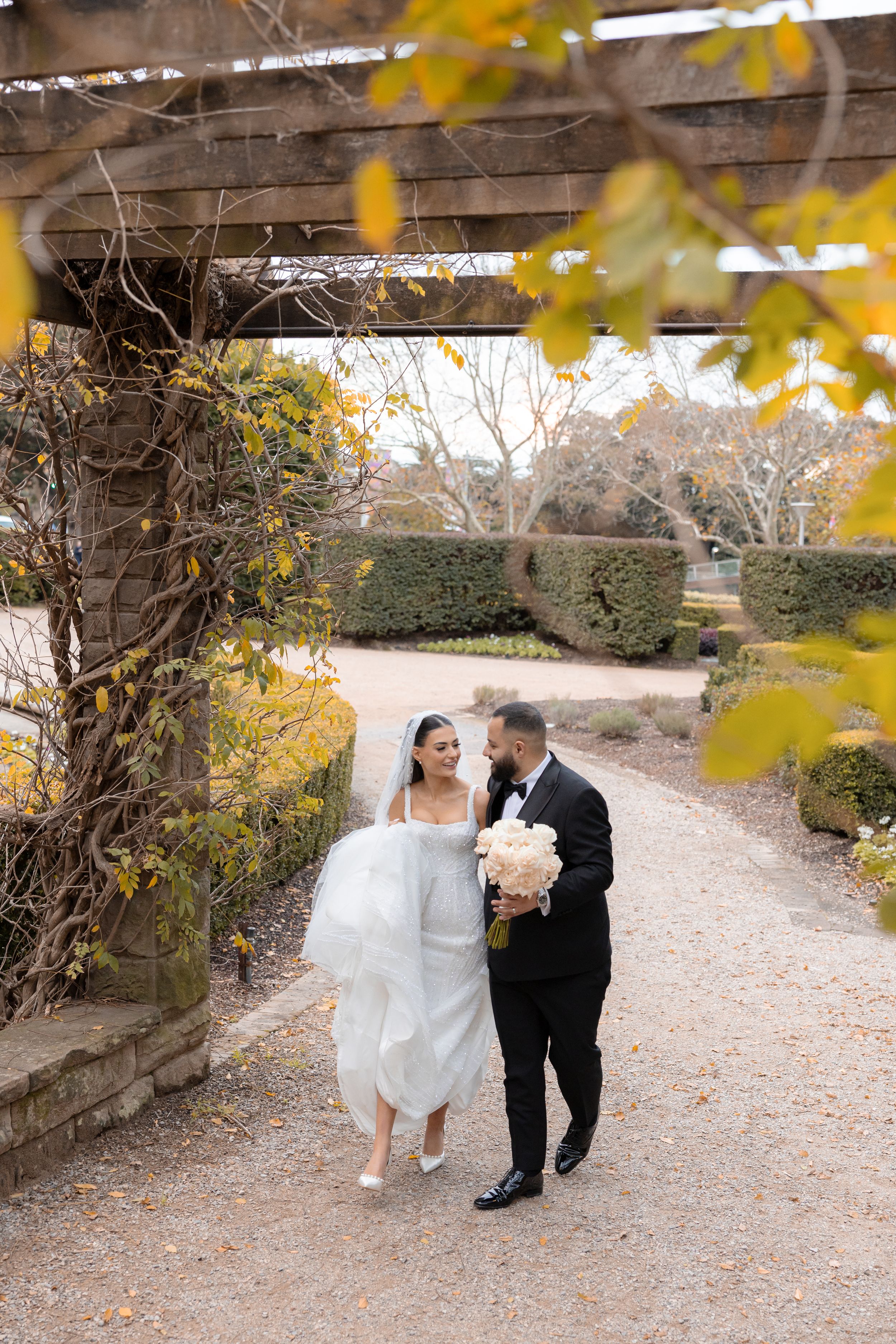 Bride and groom walking hand in hand along a garden path framed by autumn foliage and pergola.