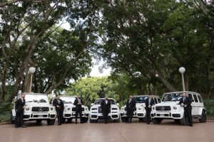 Wedding party standing beside a fleet of white luxury SUVs lined up under canopy of trees.
