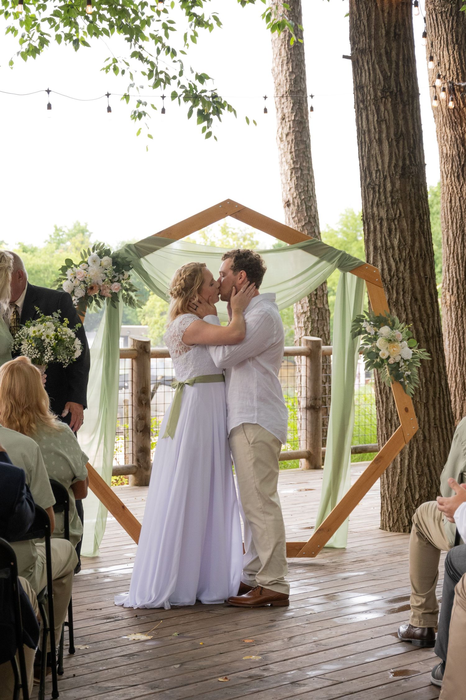 A couple shares their first kiss at a romantic woodland wedding ceremony under a hexagonal wooden arch with draped fabric.
