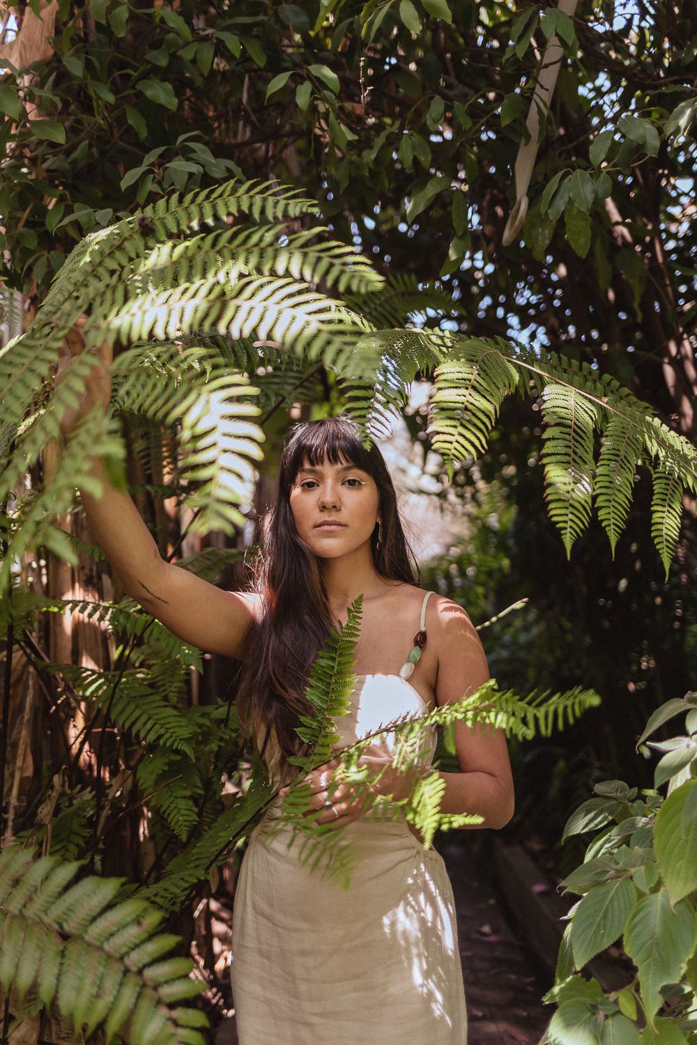 A figure stands among lush fern fronds and tropical foliage wearing a white dress in dappled sunlight.