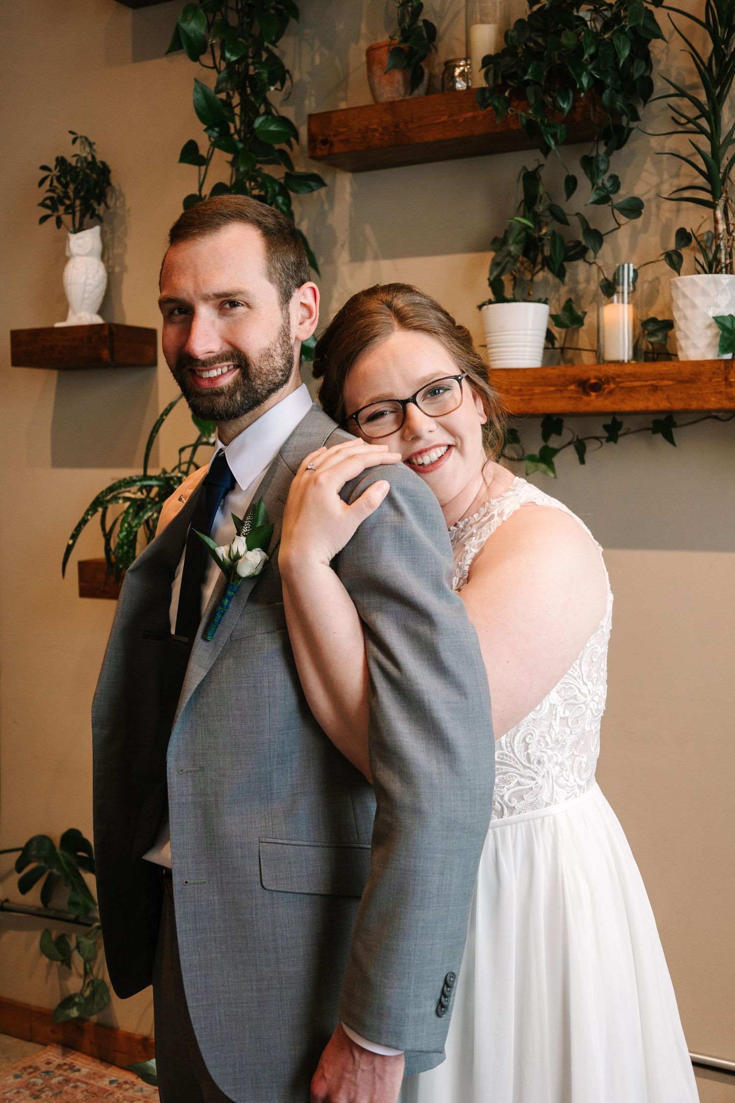 A newlywed couple poses against a wall decorated with wooden shelves and potted plants.