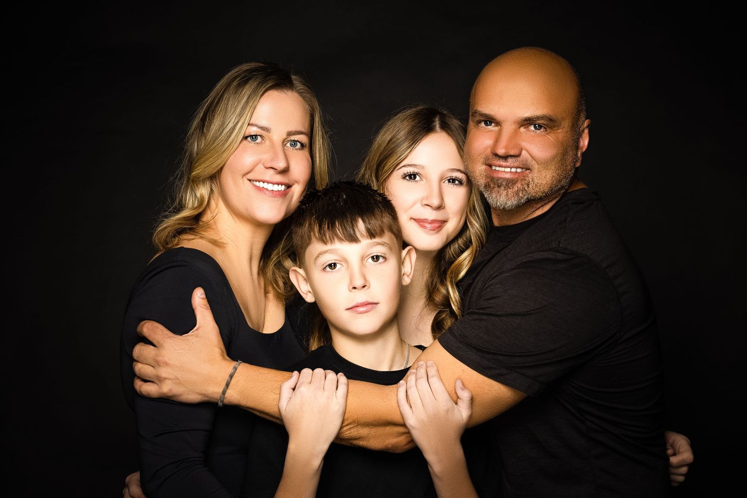 A family of four wearing dark clothes poses together for a warm portrait against a black backdrop.