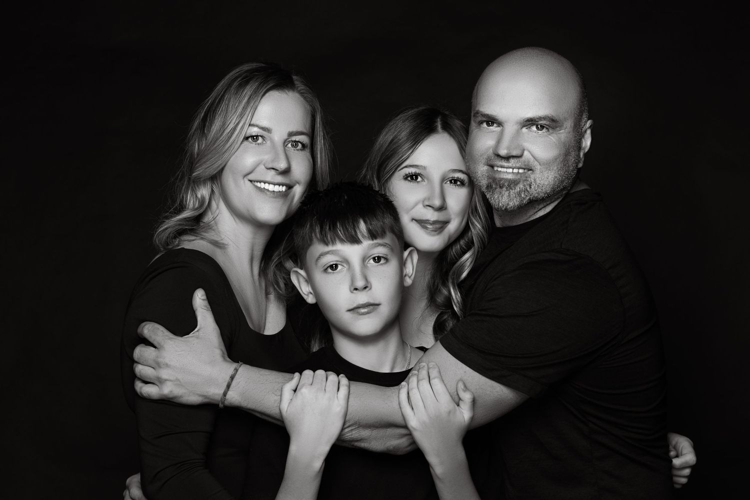 Black and white studio portrait of a family of four with dark background and natural expressions.