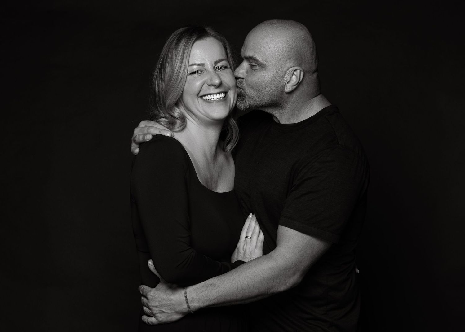 Black and white studio portrait of a couple embracing and smiling against dark background.