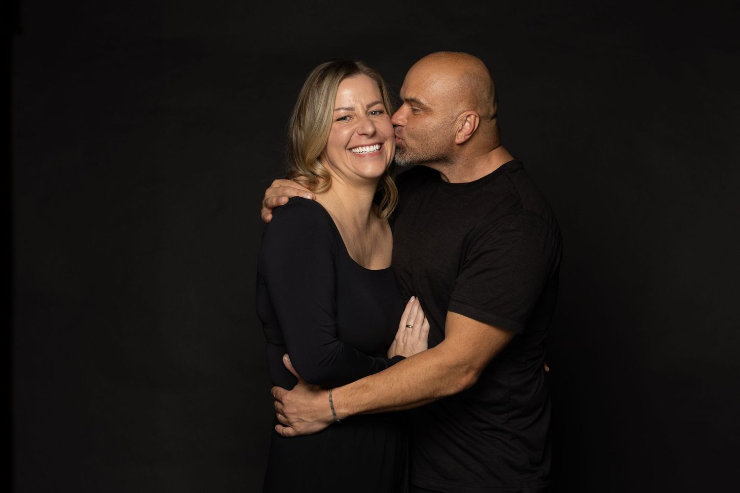 A loving couple in black tops embrace and share an intimate moment against a dark background.