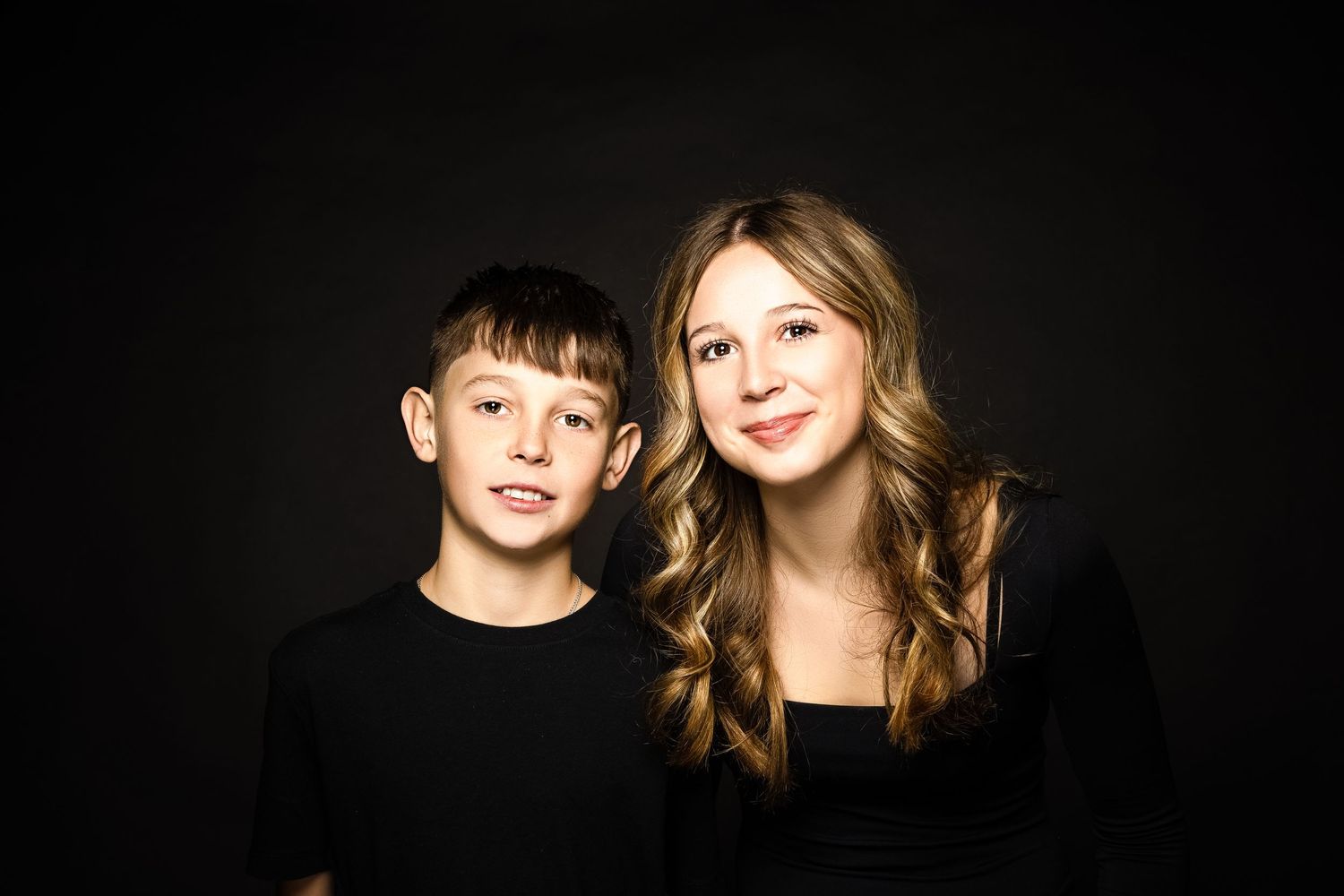A boy and girl wearing black outfits pose together against a dark background in a studio portrait.